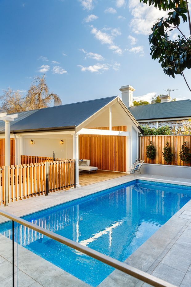 A bright residential outdoor area featuring a rectangular swimming pool surrounded by pale grey paving and glass-and-metal balustrading. A covered timber-lined pavilion or poolside lounge sits beside the pool, with white columns, a pitched dark metal roof, built-in seating and warm vertical timber screening. Timber fencing, planting, chimneys and blue sky complete the scene.