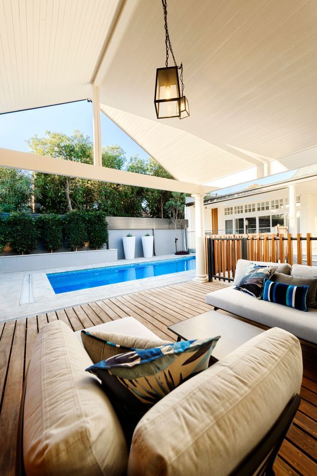 A bright covered outdoor entertaining area beside a narrow rectangular swimming pool. The space features a timber decking floor, white-painted posts and columns, a pitched white-lined ceiling, a suspended black lantern, and cushioned outdoor seating with patterned accent pillows. A timber slat privacy screen separates the seating area from the adjacent house, while lush planted screening surrounds the pool.