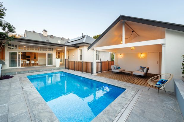 A renovated residential courtyard viewed from the pool area, featuring a bright blue swimming pool surrounded by large-format grey paving, a timber deck and covered outdoor lounge pavilion. The pavilion has a steep gabled roof with exposed timber soffit, white walls, warm wall lighting and casual seating. Glazed doors connect the outdoor area to the house, with timber privacy screening and a secondary tiled-roof structure in the background.
