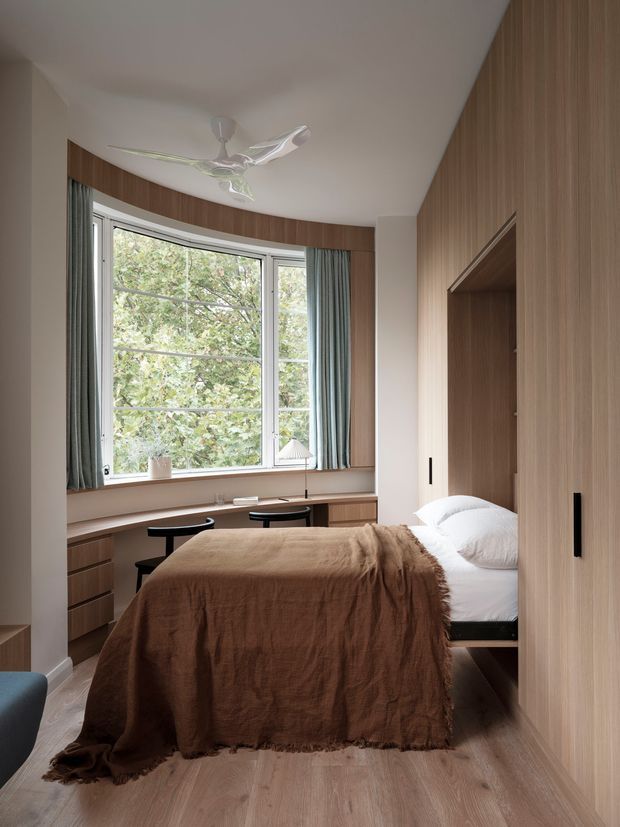 A compact, calm bedroom with a built-in wall-to-wall timber wardrobe and concealed bed nook. The room features a curved bay window with pale curtains, integrated timber cabinetry and desk seating beneath the window, light timber flooring, white bedding, a warm brown textured bedcover, and a contemporary ceiling fan. The design makes efficient use of space with streamlined built-in joinery and soft natural light.