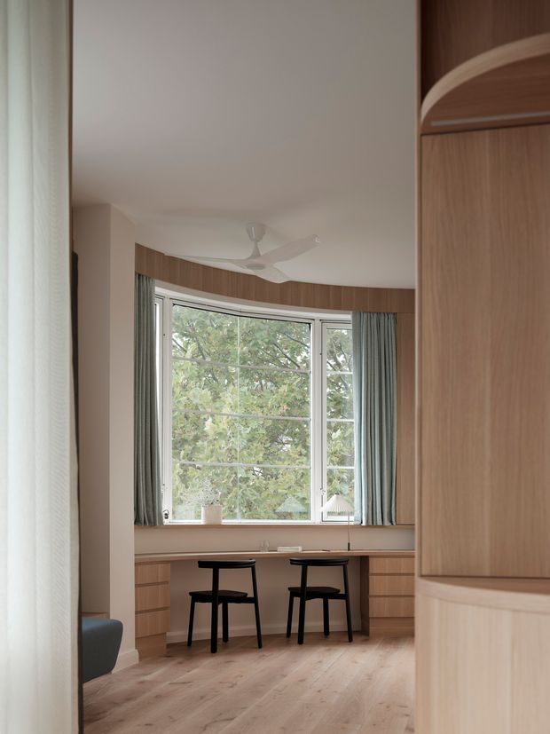 A warm, compact study or home office viewed through an opening, with a curved wall of large windows, pale blue-green curtains, built-in timber desk cabinetry, two black chairs, light timber flooring, and a white ceiling fan. A substantial timber built-in cabinet frames the right foreground.