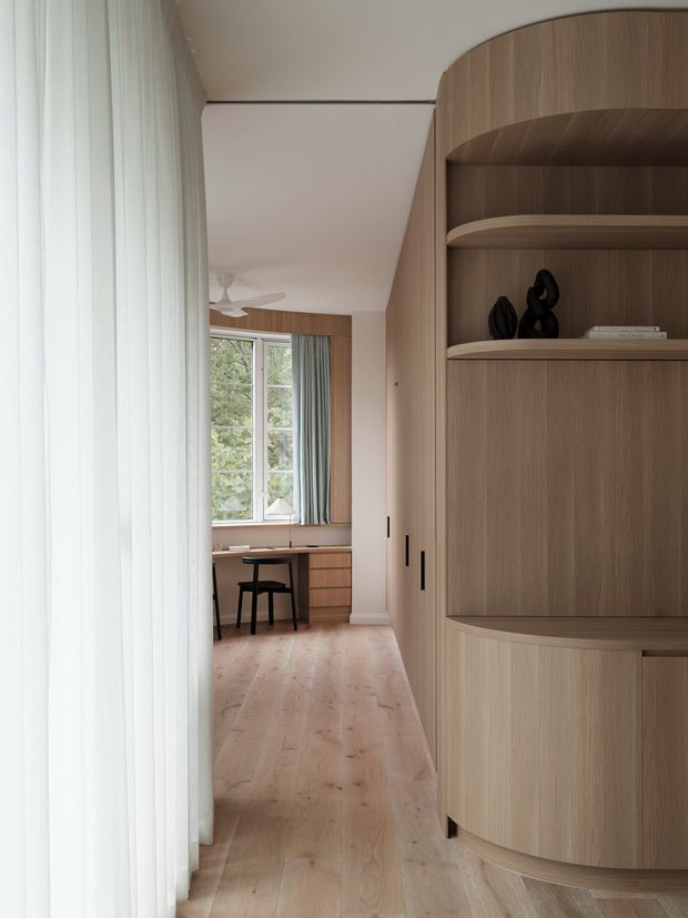 A calm, light-filled residential interior viewed along a narrow passage, with a large curved built-in timber storage and display wall on the right. The joinery has rounded edges, open shelving, concealed cabinetry and slim dark handles. Pale timber flooring continues toward a compact desk beneath tall windows, with sheer curtains and muted drapery. The room has a clean, softly contemporary character with warm natural wood and off-white surfaces.