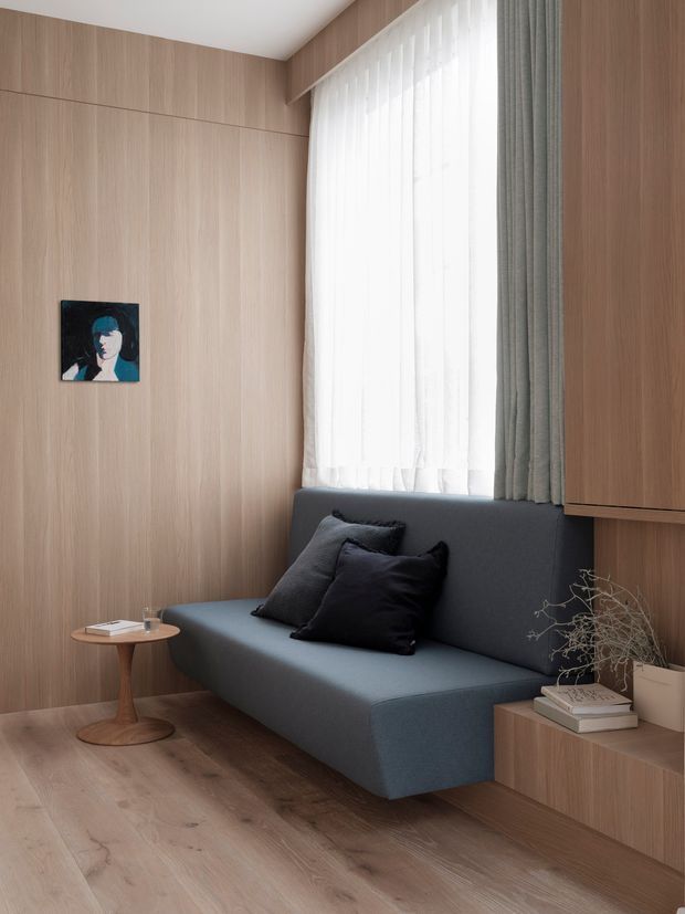 A compact, refined sitting nook with a dark blue-grey upholstered built-in sofa set against full-height light timber wall panelling. A large window with sheer white curtains and grey drapes provides soft natural light. A small round timber side table, built-in timber cabinetry, books, dried branches, and a small framed artwork complete the calm contemporary interior.