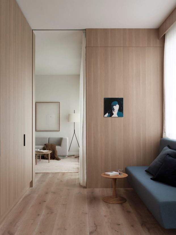 A refined minimalist interior showing a compact living room or lounge with a dark blue upholstered sofa, small timber side table, pale timber flooring, and extensive vertical timber wall panelling and built-in cabinetry. A concealed sliding door opens to an adjacent seating area with a light sofa, rug, floor lamp, and artwork. Soft daylight filters through sheer curtains, creating a calm, warm atmosphere.