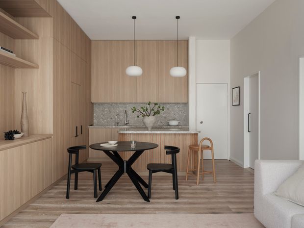 A compact open-plan kitchen and dining area with extensive light timber cabinetry and wall panelling, a grey stone-look splashback and benchtop, a small round black dining table with black chairs, and a timber bar stool at the kitchen peninsula. Two white pendant lights hang above the table. The space opens toward a partially visible living area with a pale sofa, creating a warm, refined contemporary interior.