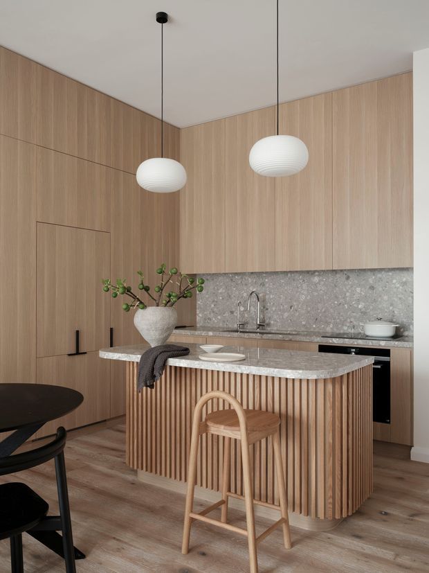 A refined contemporary kitchen with full-height warm timber wall panelling and matching integrated cabinetry. A compact island features a pale speckled stone benchtop and a vertical timber-slat front, paired with a timber bar stool. The rear benchtop has a matching stone splashback, undermount sink and minimalist black appliances. Two white ribbed pendant lights hang from the smooth ceiling, while timber flooring and restrained furnishings create a warm, calm interior.