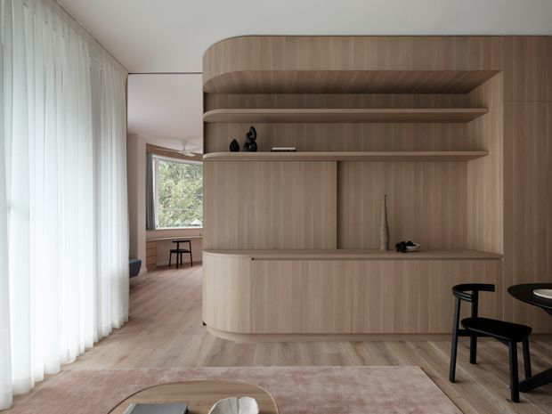 A refined open-plan living and dining area centred on a large built-in timber joinery wall with rounded corners, open shelving, concealed storage, and a recessed display niche. Pale timber flooring and sheer full-height curtains create a soft, light-filled interior, with a glimpse of a secondary seating or workspace area beyond. Black dining chairs and a low coffee table add restrained contrast.
