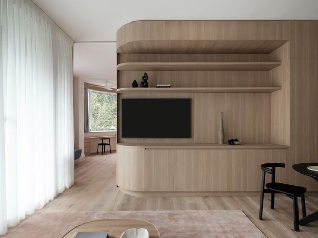 A softly lit contemporary living room centred on a large built-in timber entertainment wall with rounded corners, integrated television, open display shelving, and low cabinetry. Sheer full-height curtains line the glazed wall at left, while a timber-floored adjoining area with a small table is visible beyond. A pale upholstered seating area and rug occupy the foreground, with black dining or occasional chairs at right. The space has a warm, restrained and refined appearance.