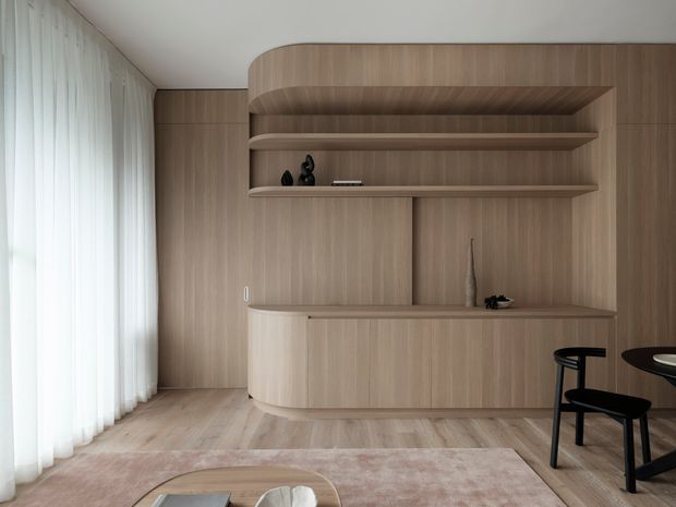 A refined contemporary living and dining area featuring extensive curved timber built-in cabinetry, open display shelving, a long integrated counter, pale timber flooring, sheer full-height curtains, and a soft neutral rug. A partially visible round dining table and black dining chairs sit to the right, while minimal decorative objects accent the joinery.