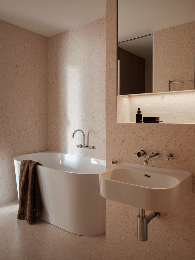 A warm, minimalist bathroom featuring a freestanding white bathtub, wall-mounted cream basin, exposed chrome plumbing, and a large mirrored recessed cabinet with integrated lighting. The walls and floor are clad in softly speckled pink-beige stone or terrazzo-like finish, creating a cohesive monochromatic interior.