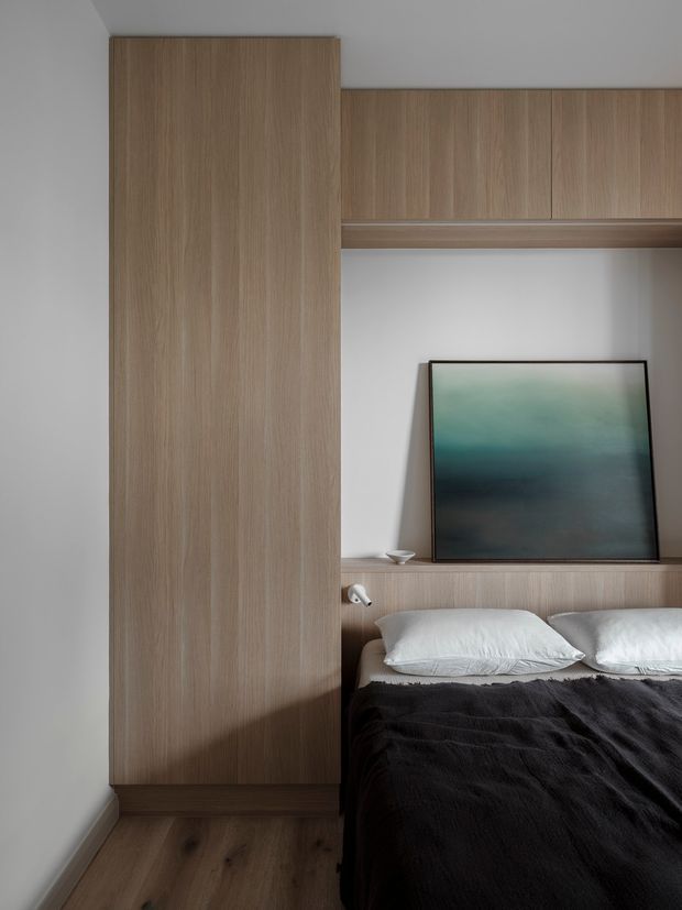 A compact contemporary bedroom with built-in light timber cabinetry spanning the wall around the bed and a recessed display niche. The room features a low platform bed with white pillows and dark bedding, a framed abstract artwork, warm timber flooring, and soft neutral walls. The joinery creates an integrated wardrobe and headboard arrangement with a clean, minimalist appearance.