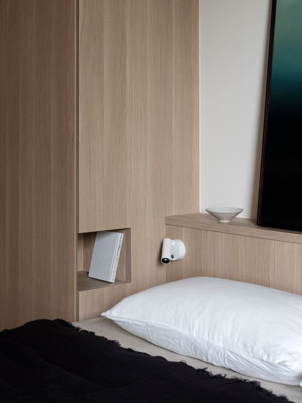 Close-up view of a minimalist bedroom corner with a bed, white pillow, black throw, and extensive light wood built-in wall cabinetry. The joinery incorporates an open book niche and a shallow shelf behind the bed, with a small wall-mounted reading light and a ceramic bowl. The edge of a black television is visible at right. The design is clean, warm, and highly integrated, with vertical timber grain and restrained neutral styling.
