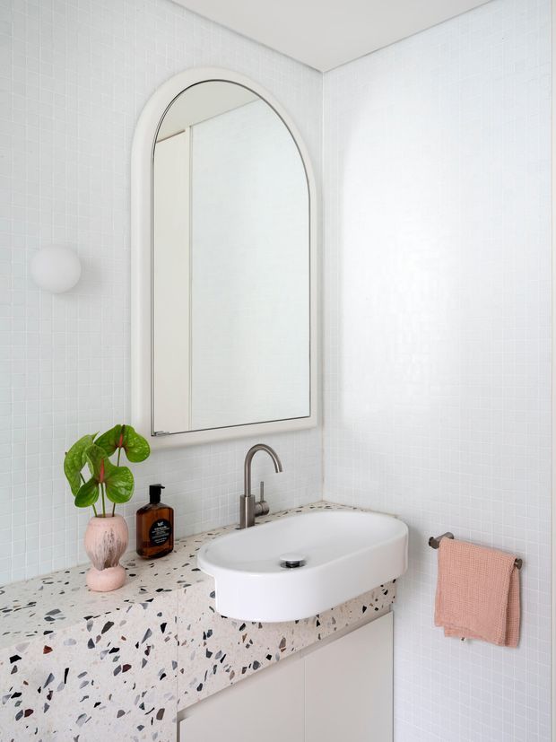 Bright, compact contemporary bathroom featuring white small-format tiled walls, a rounded-arch framed mirror, a sculptural white vessel basin with brushed metal tapware, and a terrazzo vanity benchtop with pale cabinetry. A small plant, wall sconce, and blush-pink hand towel add soft accents.