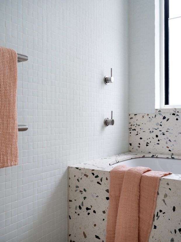 Bright contemporary bathroom featuring a terrazzo-clad bathtub and bath surround, pale small-format tiled walls, brushed metal controls, and soft peach towels. Natural light enters through the tall window at right.