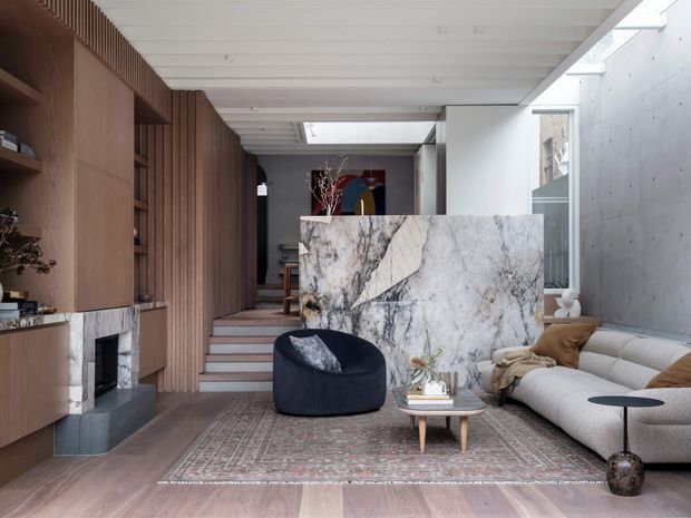 A refined contemporary living room with a large pale marble feature partition, floor-to-ceiling timber cabinetry and vertical timber battens, a concrete side wall, and a white linear ceiling with skylights. The room contains a low neutral sectional sofa, dark lounge chair, patterned rug, fireplace, and sculptural coffee tables. Steps lead toward a darker rear area, while glazing and skylights bring in natural light.