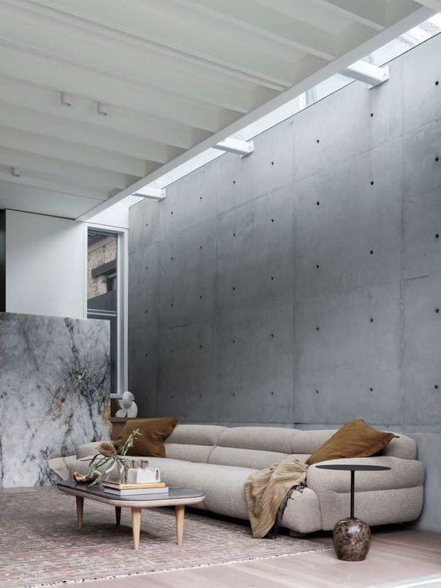 Contemporary living room with a large curved cream sectional sofa, sculptural coffee table, patterned rug, and neutral furnishings. The space features a raw concrete feature wall, white exposed ceiling beams, a narrow skylight, and a dramatic veined stone partition, creating a refined industrial atmosphere.