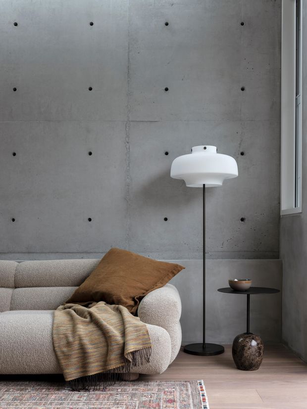 A softly styled contemporary living room featuring a pale boucle-style sofa, brown cushion, textured striped throw, sculptural white floor lamp, small black side table, and decorative vessel. The room has a raw grey concrete feature wall with regular circular form-tie marks, a narrow window, pale timber flooring, and a patterned rug.