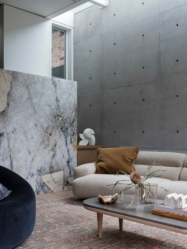 A refined contemporary living room with a curved cream boucle sofa, dark rounded lounge chair, low timber coffee table, patterned rug, sculptural decor, and a large veined stone slab partition. The room features a tall raw concrete feature wall with visible form-tie marks, a narrow window, and a skylight, creating a sophisticated material-focused interior.