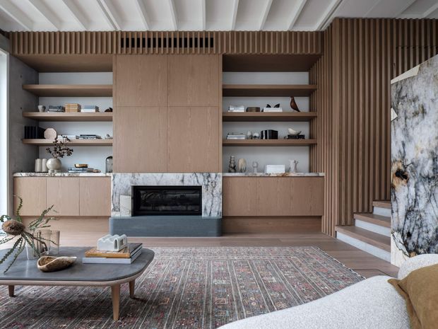 A refined contemporary living room centred on a built-in timber media and fireplace wall. Open shelving, pale timber cabinetry, a dark fireplace surround, veined stone accents, vertical timber battens, a white-beamed ceiling, and a patterned area rug create a warm, layered interior. A staircase rises at the right, while a low coffee table and upholstered seating occupy the foreground.