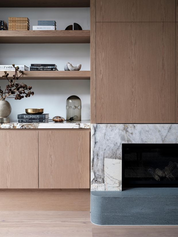 A close-up of a refined living room built-in joinery and fireplace. Warm light-oak cabinetry and open shelving display books and decorative objects, while a large veined marble surround frames a black fireplace with a dark stone hearth. Pale timber flooring and restrained contemporary styling create a calm, sophisticated composition.