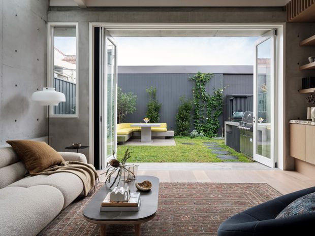 A contemporary living room opening directly onto a private landscaped courtyard through wide bi-fold doors. The room features a textured concrete wall, pale upholstered seating, a dark coffee table, patterned rug, built-in timber cabinetry and open shelving. Outside, a lawn, dark vertical fence, paved path, climbing greenery, outdoor seating and a built-in barbecue are visible.