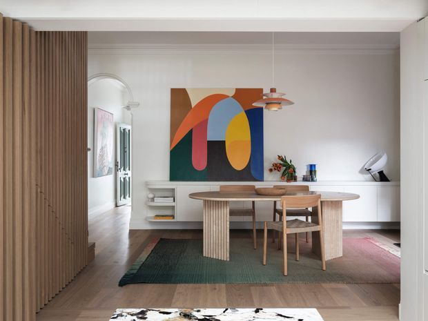 A refined open-plan dining area with a sculptural timber dining table and chairs, set on layered rugs over a warm timber floor. Vertical timber battens form a partition on the left, while white built-in cabinetry lines the rear wall. A large colourful abstract artwork provides a bold focal point, complemented by contemporary pendant lighting and restrained decorative objects. An arched opening connects the dining area to an adjacent space.