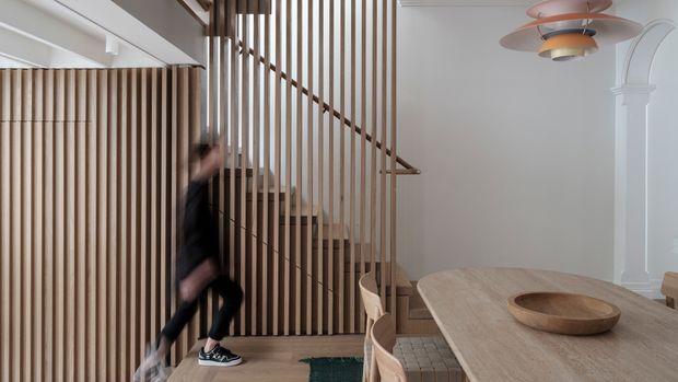 Bright contemporary interior featuring a timber staircase screened by vertical wood battens, with matching timber wall panelling, a light wood dining table, white walls, and a sculptural layered pendant light. A blurred person is moving on the stairs.
