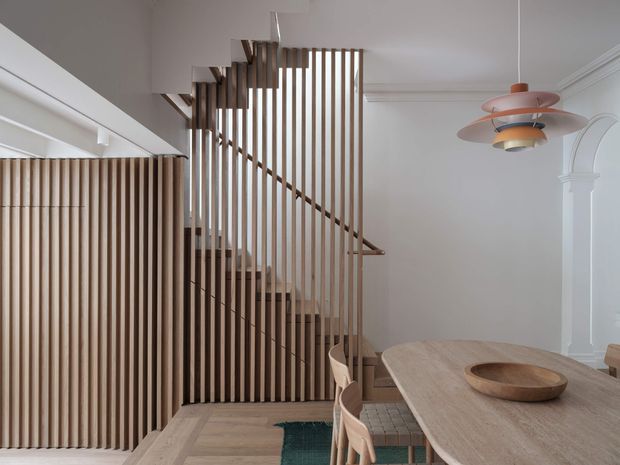 A refined open-plan dining and stair area with a light timber dining table and chairs in the foreground. A sculptural timber staircase is screened by evenly spaced vertical timber slats, creating a strong architectural feature and partial separation from the adjacent area. The room has white walls with traditional moulded detailing and an arched wall opening, pale timber flooring, and a layered copper-toned pendant light.