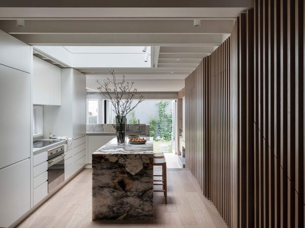 A refined contemporary kitchen viewed from the entry, featuring white full-height cabinetry, integrated appliances, a dramatic dark-veined stone island with timber stools, pale timber flooring, and a vertical timber slat screen along the right wall. Exposed ceiling beams and a linear skylight bring natural light into the space, with a small planted courtyard visible beyond.