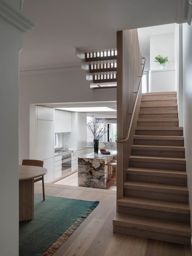 Contemporary interior view from a hallway or landing, featuring a sculptural timber staircase with vertical slatted balustrade and integrated handrail. A bright white kitchen with a large veined stone island is visible beyond, while a small dining area and muted teal rug occupy the foreground. The space combines pale timber flooring, white walls and cabinetry, and refined minimalist detailing.