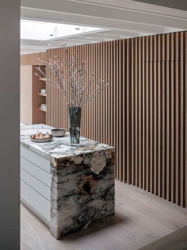 A refined contemporary kitchen featuring a compact island with a dramatic veined natural stone waterfall benchtop and white drawer cabinetry. A full-height vertical timber slat wall provides a warm architectural feature, with integrated shelving and a pale timber floor. The ceiling has white recessed panel detailing and a skylight or clerestory opening. Minimal decorative styling includes a dark glass vase with branches and ceramic bowls.