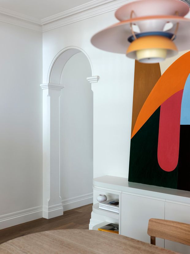 Bright, refined interior corner with white decorative wall mouldings and a recessed arched niche, dark timber flooring, and a low white built-in media cabinet. A colourful abstract artwork and sculptural layered pendant light provide bold focal points, with part of a timber dining table and chair visible in the foreground.