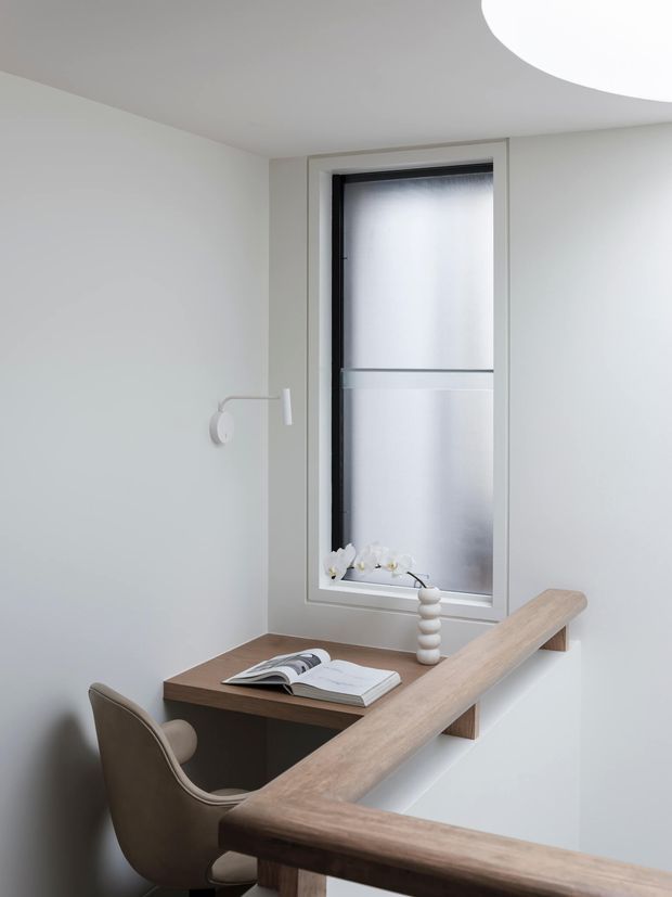 A compact, minimalist home office or study nook beside a stair or landing. The space features a built-in light wood desk and timber handrail, a sculptural upholstered chair, a frosted vertical window, pale walls, a small wall-mounted reading light, and simple decorative objects. The curved ceiling opening adds a distinctive architectural detail.