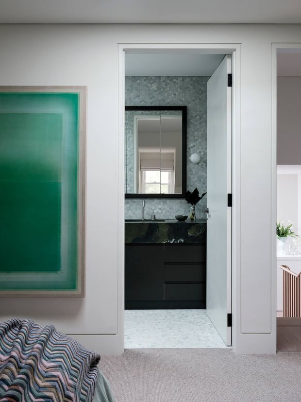 A refined bedroom view looking through an open doorway into a bathroom. The bedroom has pale walls, carpeted flooring, a large green artwork, and patterned bedding in the foreground. The bathroom features pale patterned wall and floor tiles, a dark stone benchtop, black cabinetry, a wall mirror, and white fixtures. A glimpse of an adjacent bright room is visible at right.