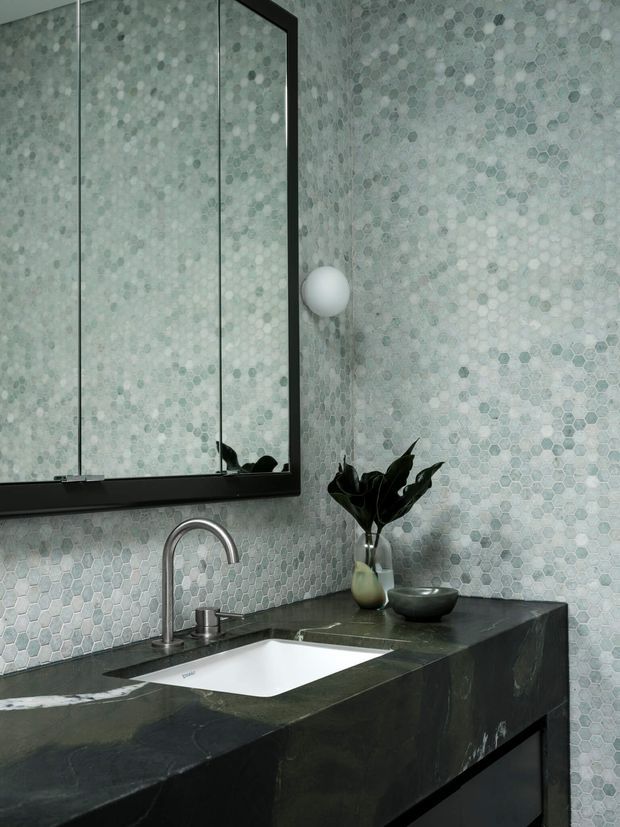 Close-up of a refined bathroom vanity with a dark stone benchtop and integrated white rectangular basin, brushed metal gooseneck tap, black-framed mirror, and pale grey hexagonal mosaic wall tiles. A small plant and bowl provide restrained decorative accents.