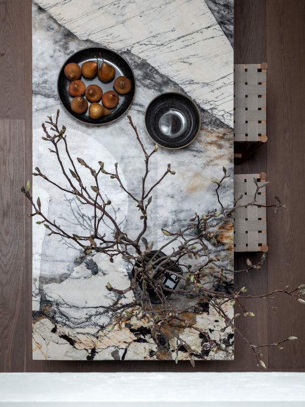 Top-down view of a refined dining table setting. A large dramatic veined stone slab forms the tabletop or central table surface, surrounded by dark timber flooring or table framing. Black bowls hold fruit, while bare branches create a sculptural natural arrangement. Two woven or upholstered dining chairs are partially visible along the right side. The composition is moody, organic, and editorial.