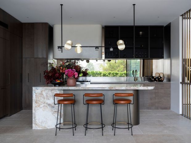A sophisticated contemporary kitchen centred on a large island with a pale veined stone waterfall benchtop and three sculptural leather-and-metal bar stools. Dark timber cabinetry spans the left wall, while black glazed upper cabinetry and a mirrored or reflective splashback sit above the rear benchtop. Large windows bring in greenery and daylight. A sculptural pendant light hangs over the island, and vertical timber screening is visible at the right edge. The palette is dark wood, black, grey stone and warm brown leather, creating a refined, moody yet naturally lit interior.