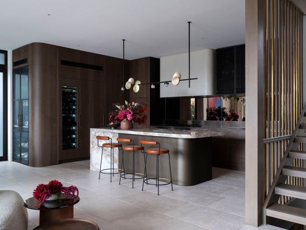 A sophisticated open-plan kitchen and living area with dark timber cabinetry, a large curved island with a light stone benchtop, and four slim bar stools. Pendant lights hang above the island, while a wine fridge and integrated appliances are built into the cabinetry. A partially screened staircase with vertical timber slats is visible at right, and large-format pale floor tiles create a refined, contemporary interior.