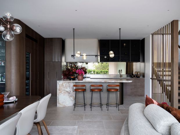 Open-plan contemporary kitchen and living area with a central marble-clad island, dark timber cabinetry, black overhead cupboards, and three bar stools. A dining area with sculptural white chairs sits to the left, while a light sofa occupies the foreground. Vertical timber screening separates the living zone, and pendant and globe lighting add decorative detail.