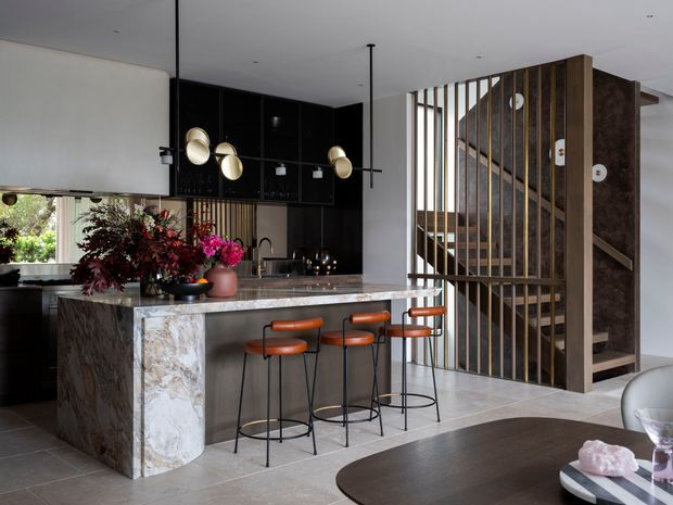 Open-plan contemporary kitchen and dining area with a large veined stone island, dark glossy cabinetry, integrated pendant lighting, and four orange-brown bar stools. A dramatic timber slatted screen encloses the stairway at right, while a round dining table occupies the foreground. The palette is dark, warm, and refined, with strong contrast between black joinery, pale stone, timber, and white walls.