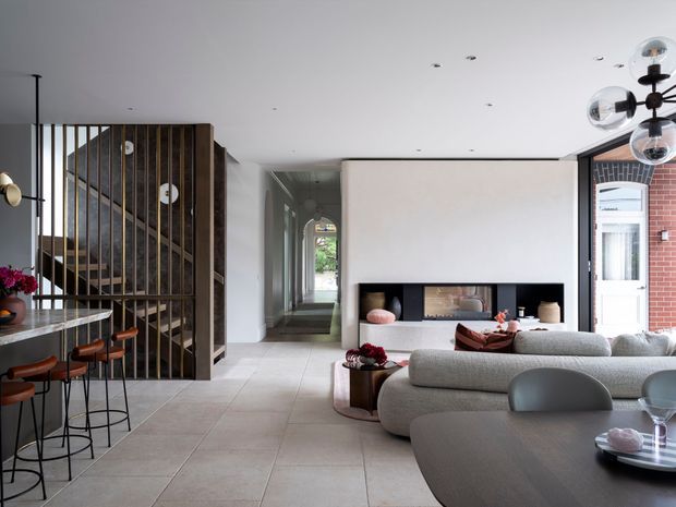 Open-plan contemporary interior combining a living room, dining area and kitchen. A sculptural curved sectional sofa faces a minimalist fireplace set into a smooth white wall, while a dark stone island with bar seating occupies the left side. A vertical timber-and-metal slatted stair screen creates a strong architectural feature, and large pale floor tiles unify the space. The room is softly lit with a modern globe pendant and recessed ceiling lights.