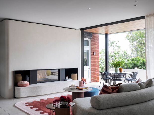 Contemporary living room with a large sculptural fireplace wall, built-in open shelving, low textured hearth, rounded grey sectional sofa, nesting coffee tables, and a soft abstract rug. Full-height glazing opens to a covered outdoor dining terrace, with exposed red brick and timber-lined soffit visible outside. The room is bright and refined with a neutral palette and warm natural accents.