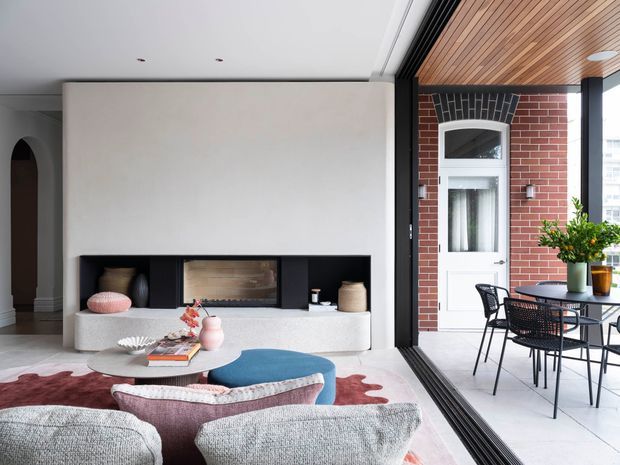 Contemporary open-plan living room opening onto a covered terrace through fully retractable glazing. The interior features a minimalist plaster fireplace wall with dark recessed shelving and a timber firebox surround, a low built-in bench, sculptural coffee table, and soft upholstered seating. The terrace continues the clean palette with a brick wall, white door, timber-lined ceiling, and black-framed outdoor dining furniture.