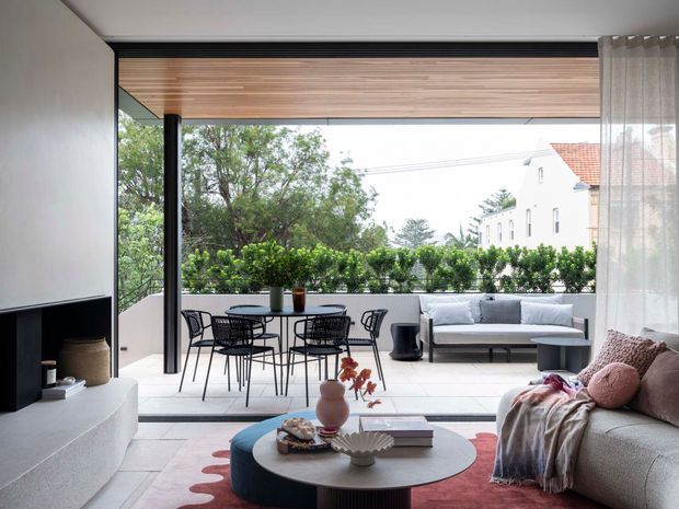 A bright contemporary living room opening onto a covered terrace through large retractable glazing. The room features a curved light sofa, round coffee table, pale tiled floor, minimalist dark fireplace and media joinery, timber-lined ceiling, sheer curtains, and a small outdoor dining and lounge arrangement overlooking greenery.