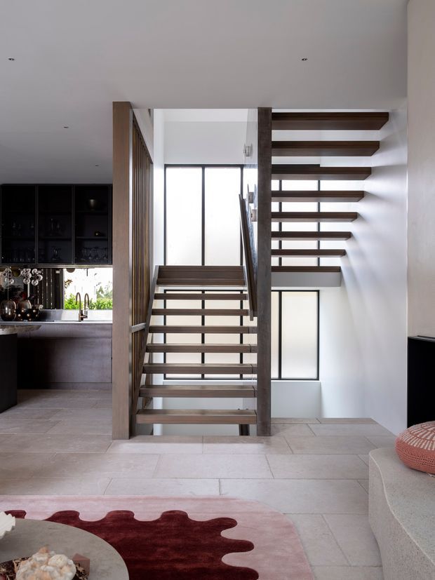 Contemporary open-plan interior focused on a floating open-riser staircase with dark timber treads, vertical timber screening, and substantial timber posts. The stair connects a bright upper level to the living area below. A dark, highly fitted kitchen with display shelving is visible to the left, while pale tiled flooring, white walls, large windows, and a partial curved sofa create a clean, light-filled setting. A sculptural red-and-pink rug adds colour in the foreground.