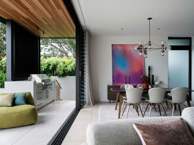 Contemporary open-plan dining and living area connected to a covered outdoor kitchen through fully retractable glazing. The interior features a large abstract artwork, sculptural multi-globe pendant light, dark timber dining sideboard, white upholstered dining chairs, and pale neutral finishes. A green sofa and additional seating occupy the foreground, while the outdoor area includes built-in stainless-steel barbecue cabinetry beneath a warm timber-lined ceiling.