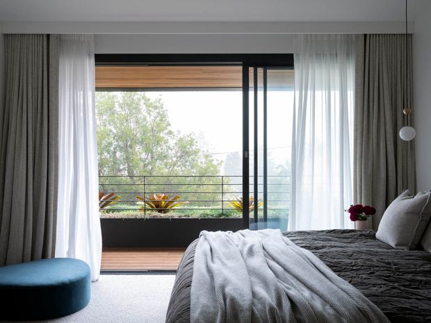 A contemporary bedroom with a large bed dressed in dark grey bedding and a light grey throw, framed by full-height sheer and heavy grey curtains. Black-framed sliding glazing opens onto a timber-floored balcony with a simple black railing and planted greenery beyond. A blue upholstered ottoman and a minimal wall-mounted globe light add restrained accents.