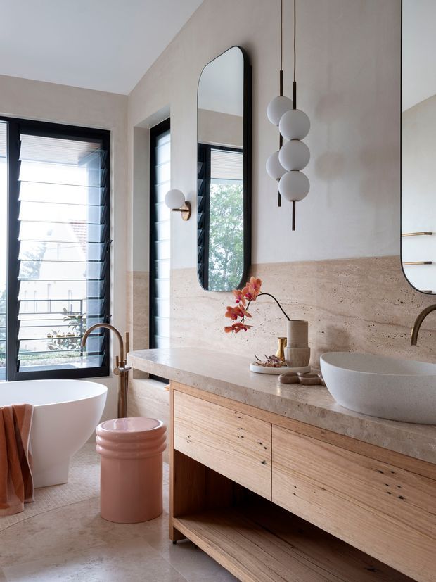 A refined contemporary bathroom featuring a freestanding white bathtub, a sculptural white vessel basin, and a long floating timber vanity with integrated storage. The walls and benchtop are clad in warm beige natural stone, complemented by black-framed louvre windows, tall rounded-edge mirrors, and a cluster of spherical pendant lights. Soft daylight, light timber, and muted pink accents create a warm, luxurious atmosphere.