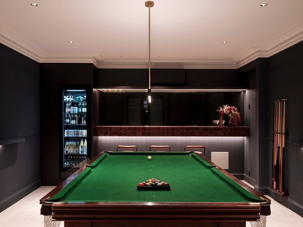A dark, refined billiards room centered on a full-size green-felt pool table. The room features deep charcoal wall panelling, dark timber cabinetry, a back bar with illuminated shelving and drinks refrigerator, a slim pendant light above the table, recessed ceiling lights, decorative crown moulding, cue storage, and burgundy-toned bar detailing.