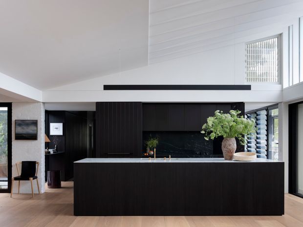 A contemporary open-plan kitchen with a large dark wood island and matching full-height cabinetry, contrasted by a pale stone benchtop and dark splashback. The room has light timber flooring, white horizontal wall panelling, extensive glazing and louvred windows, and a high ceiling with a slim black linear pendant. A small adjacent sitting area is visible at left.